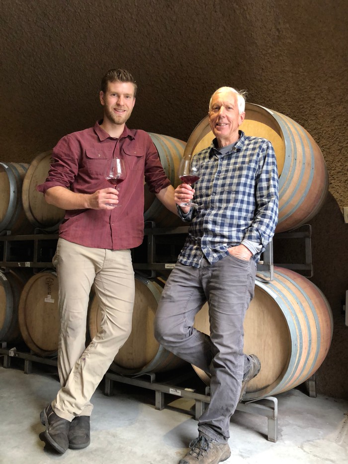 2 men holding wine glasses in front of wine barrels